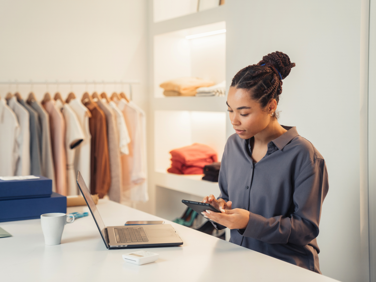 A retail worker stands at a counter using both a laptop and smartphone in a modern boutique, with clothes hanging on racks in the background.