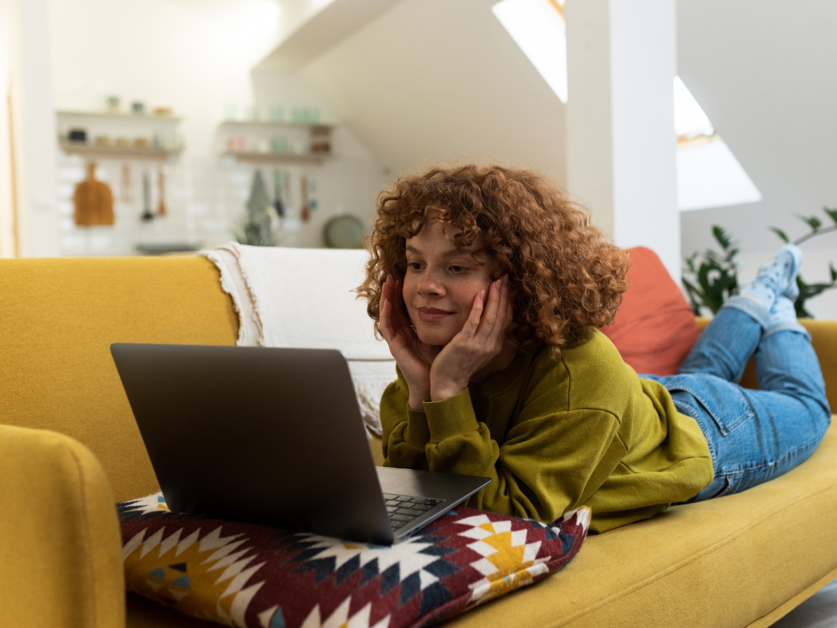 A woman with curly hair is lying on a yellow couch, using a laptop. She appears relaxed and focused, with her hands supporting her face, in a cozy indoor setting.