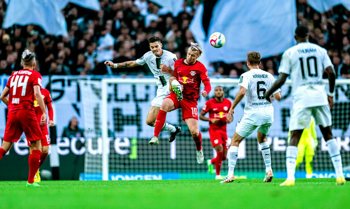 Soccer players from opposing teams in red and white jerseys compete for the ball mid-air during a match, with a crowd in the background.