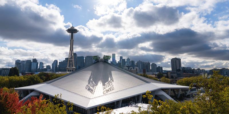 Seattle skyline featuring the Space Needle, with Climate Pledge Arena in the foreground surrounded by autumn trees under a partly cloudy sky.
