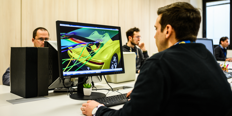A person working on a computer displaying a 3D design of a yellow car in an office setting.