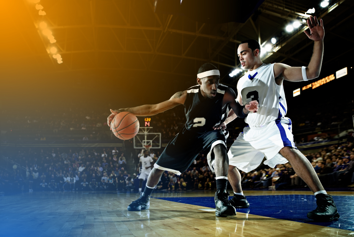 Two basketball players compete on the court in an intense game, with one player dribbling the ball while being defended by an opponent. The scene takes place in a packed arena with visible hoop and scoreboard, under bright stadium lights.