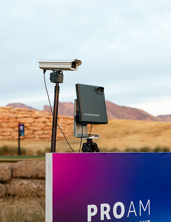 A TrackMan device and video camera set up at a golf course with a sign reading 'PRO AM', likely for advanced shot tracking or event analytics at a PGA Tour event.