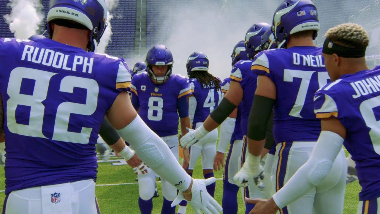 Players from the Vikings football team make a dramatic entrance onto the field, running through smoke and giving high fives to teammates while dressed in purple uniforms.