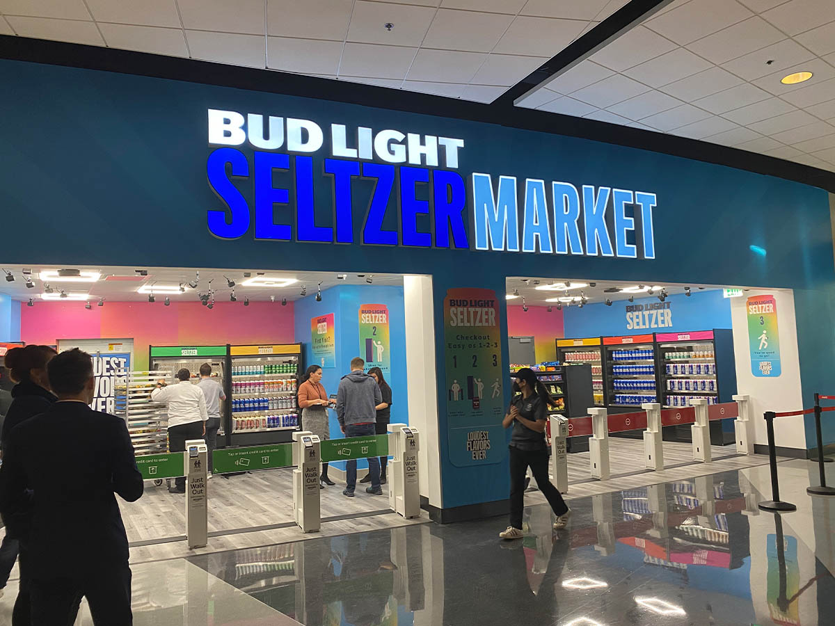The entrance to a Bud Light Seltzer Market grab-and-go store featuring prominent branding, open entry gates, and customers selecting beverages inside the brightly lit and colorful store.