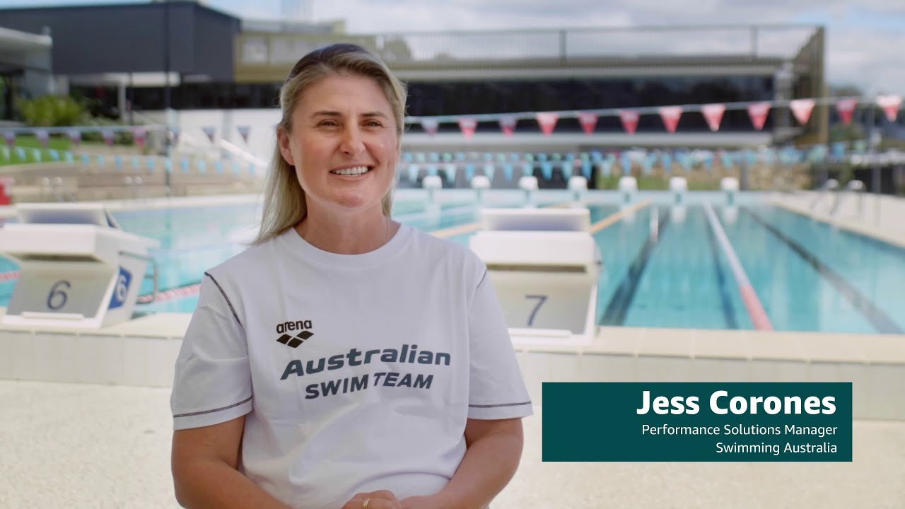 Jess Corones, Performance Solutions Manager for Swimming Australia, is being interviewed by a swimming pool, wearing an Australian Swim Team shirt. The background features swimming lanes and diving blocks, indicating a professional aquatic setting.