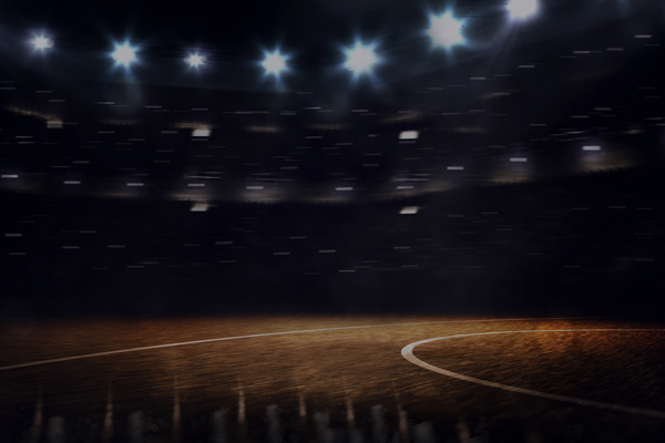 A wide view of an empty, illuminated basketball court inside a large stadium. The arena lights are on, and the stands are dimly visible in the background, evoking the atmosphere before or after a game.