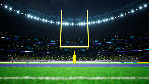 A nighttime view of an NFL football stadium, featuring a brightly lit goalpost and green field with stadium lights illuminating the stands filled with spectators.