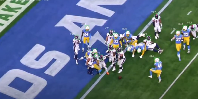 Football players from two teams scramble near the end zone during a game, with the ball on the ground.