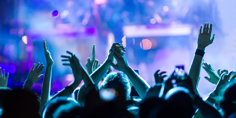 A crowd of people raising their hands at a concert, illuminated by vibrant blue and purple stage lights.