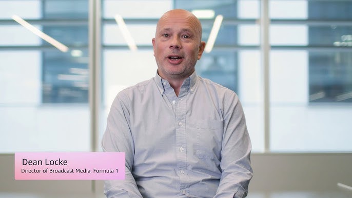 A person in a light blue shirt sits in a modern office setting with large windows; a caption identifies them as Dean Locke, Director of Broadcast Media, Formula 1.