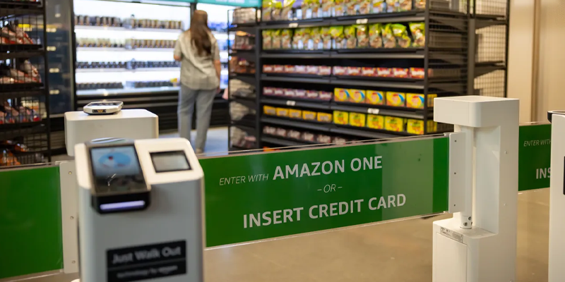Entrance to an Amazon Go store with a sign reading "Enter with Amazon One or Insert Credit Card," and shelves of snacks visible in the background.