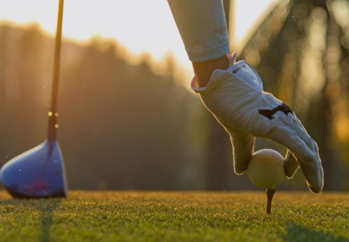A golfer's gloved hand placing a golf ball on a tee at sunrise, with a golf club nearby.
