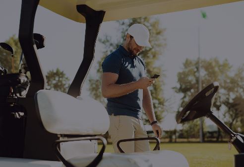 A person standing next to a golf cart, looking at a smartphone, with a sunny outdoor setting and trees in the background.
