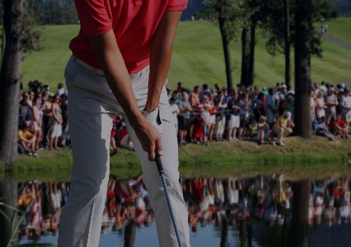 Golfer in a red shirt preparing to putt near a water hazard, with a crowd of spectators in the background.