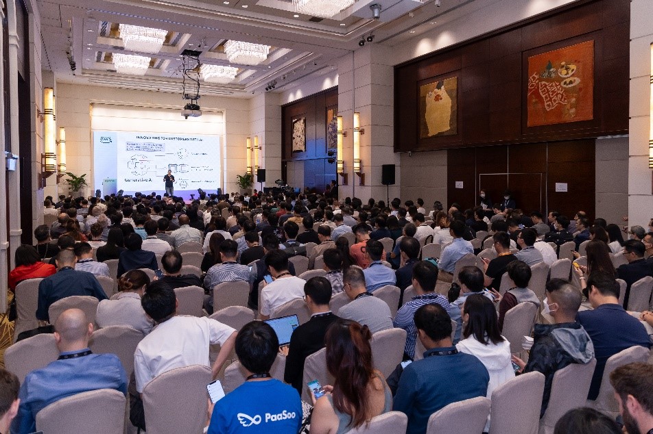 A large conference audience attends an AWS Startup Day presentation in a well-lit hall with a speaker presenting slides on stage.
