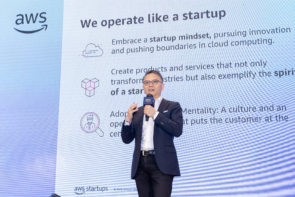A speaker presents on stage at the AWS Startup Summit with a large screen in the background that reads 'We operate like a startup,' highlighting topics such as startup mindset, cloud computing innovation, and customer-centric culture. The event emphasizes startup principles in cloud computing and business transformation.