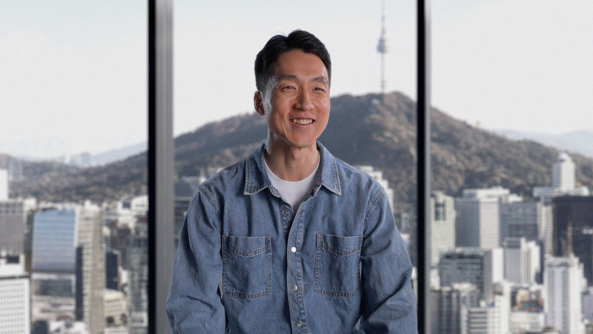 Minyong Lee, Founder and CEO of Grey Box, in a denim shirt by a window with a Seoul cityscape