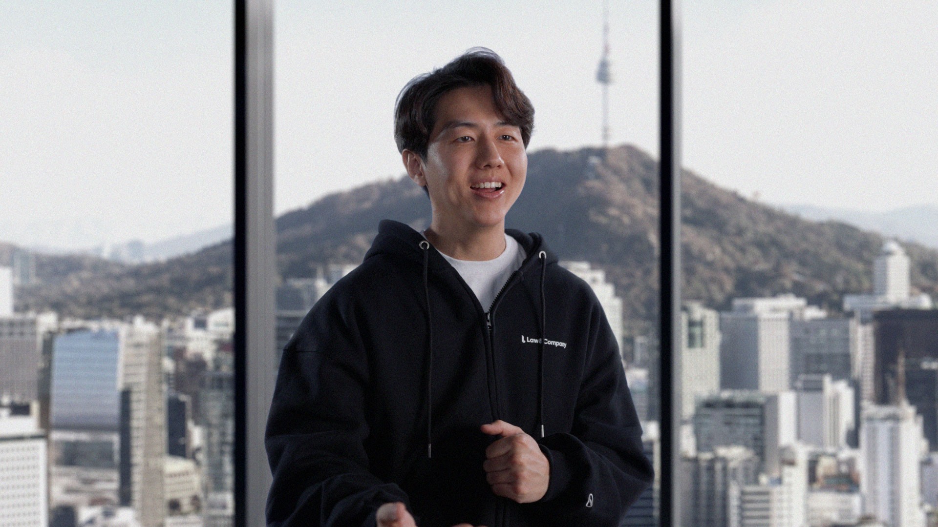 A Law and Company founder in a black hoodie in front of a Seoul city view through a window
