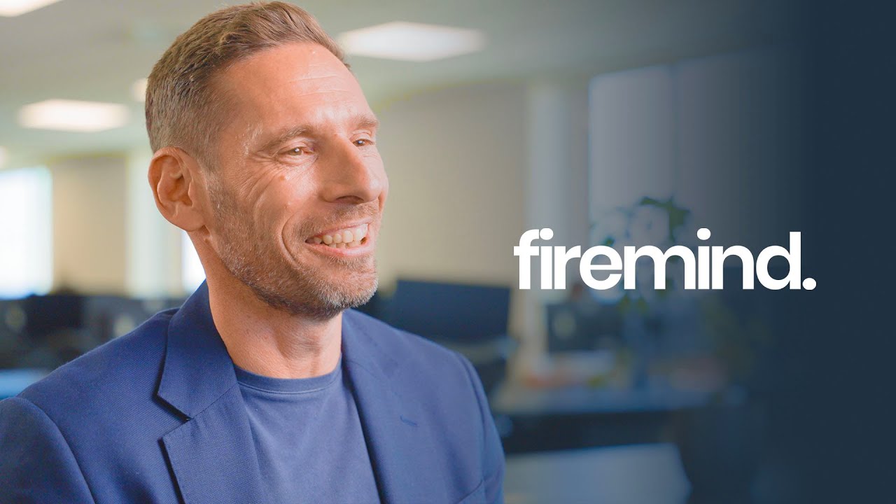 A smiling man in a blue blazer is shown in a well-lit office environment with the word 'firemind.' prominently displayed on the right side, serving as a YouTube thumbnail for a Firemind interview video.