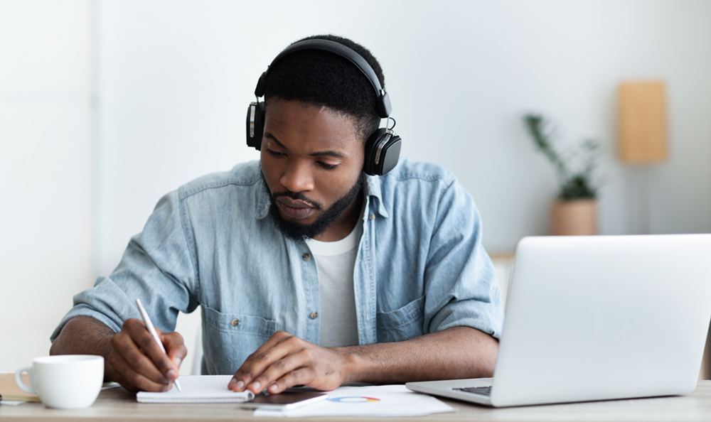 A man wearing headphones is studying at a desk with a notebook and an open laptop in front of him. He is writing notes and appears focused on his work.