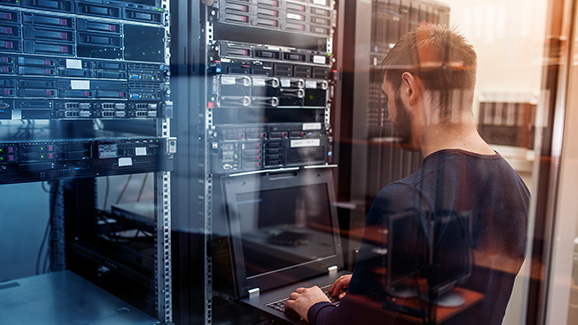 An IT professional is working on a laptop in a server room, surrounded by network servers and equipment. The image is viewed through glass, reflecting a technology-focused and secure infrastructure environment.