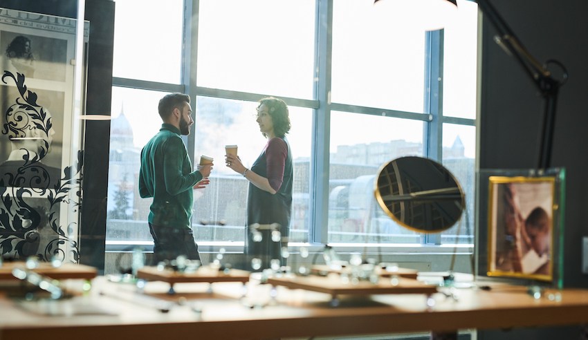 Two colleagues have a conversation while holding coffee cups near a large window in a modern office space, with a desk and office items in the foreground.