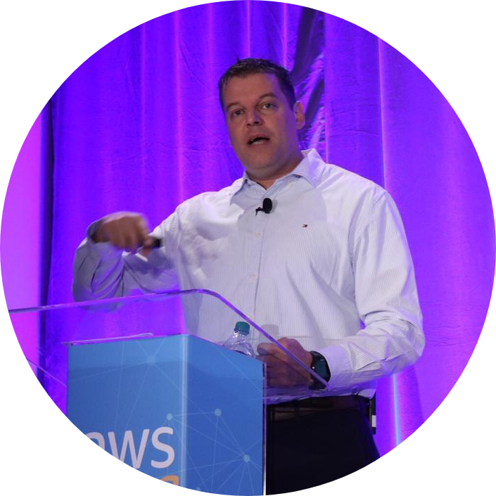 A speaker presenting at an AWS conference, standing behind a podium with an AWS logo, in front of a purple background.