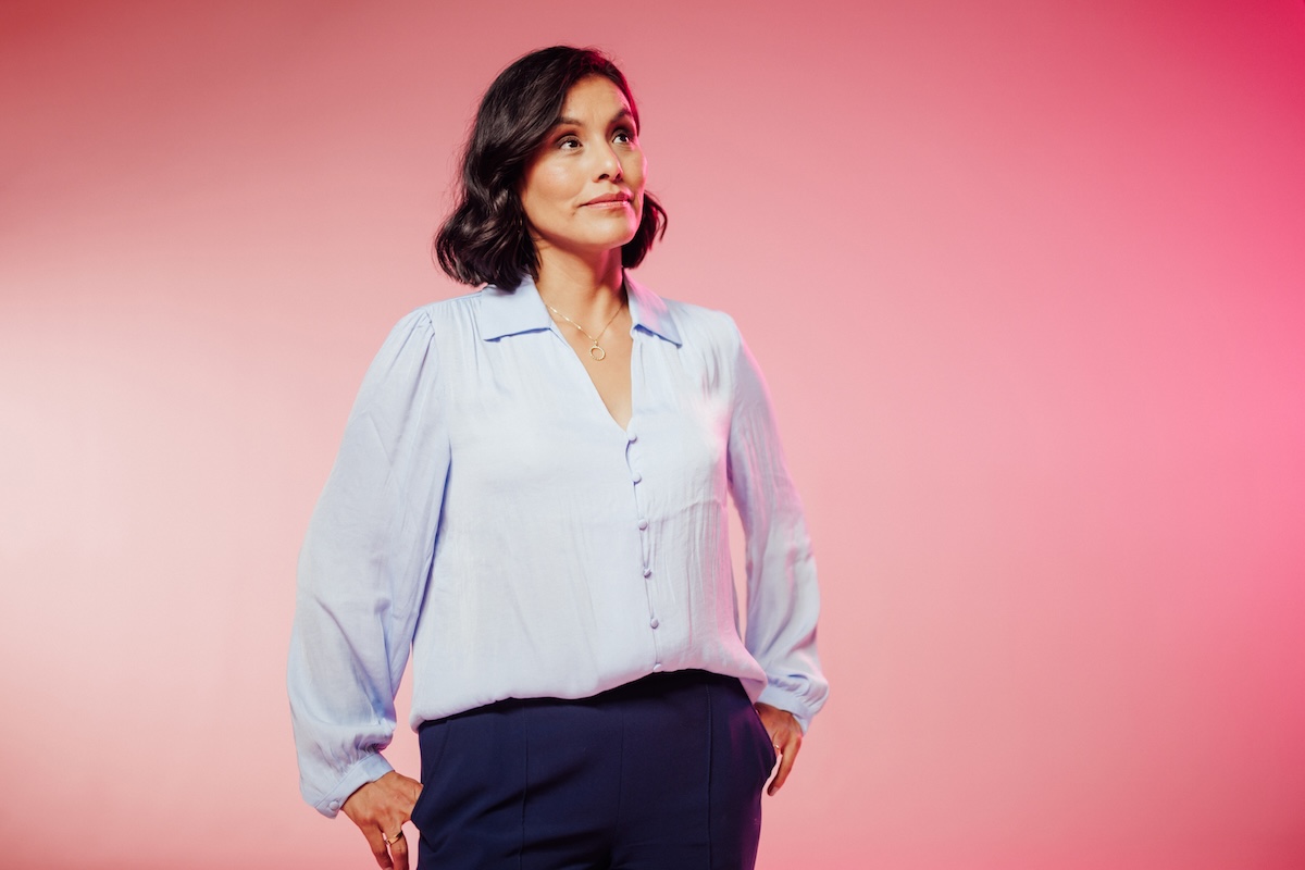 Portrait of a woman with short dark hair wearing a light blue blouse and dark pants, standing with hands in pockets against a pink background.