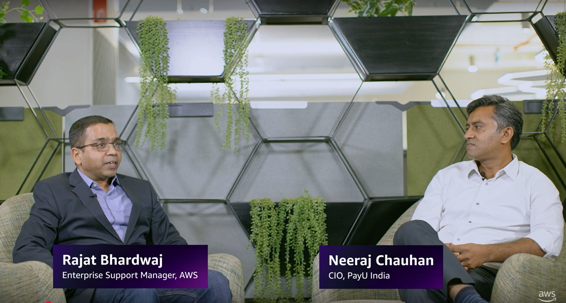 Rajat Bhardwaj from AWS and Neeraj Chauhan from PayU India having a conversation in a modern office with hexagonal shelves and green plants in the background.