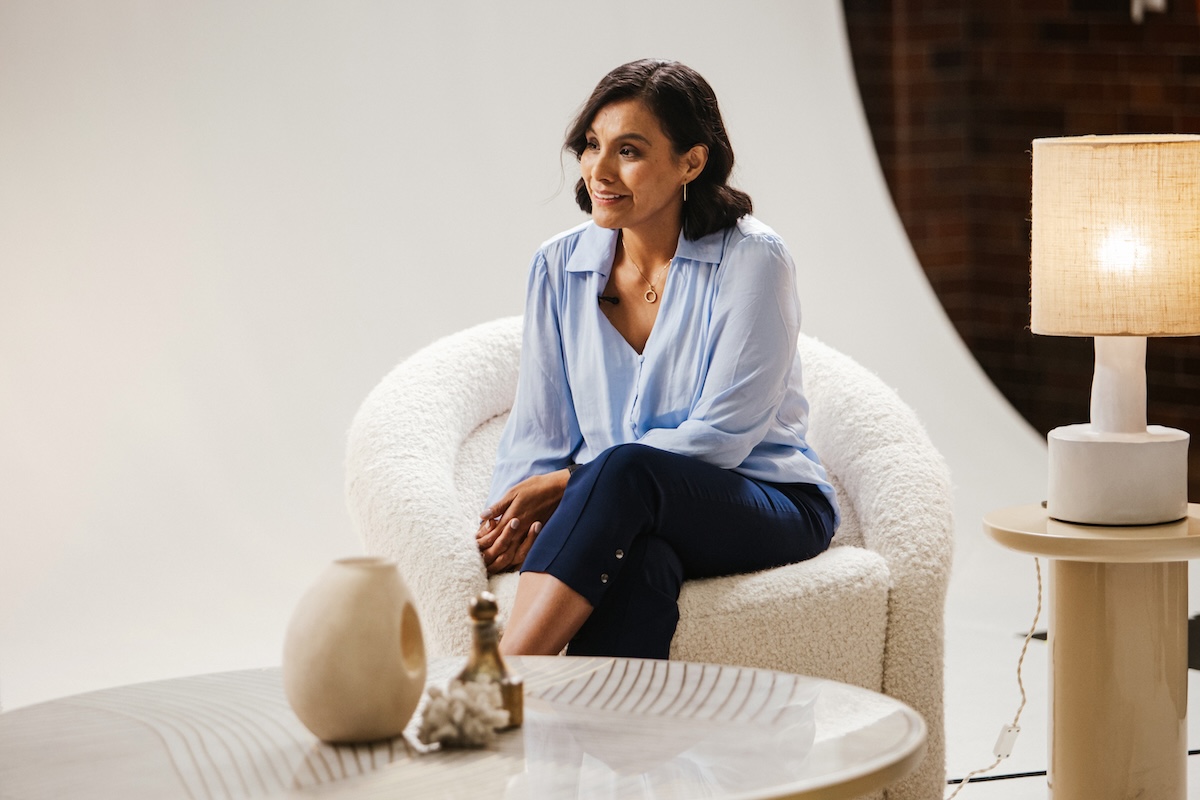 A woman wearing a light blue blouse and dark pants is seated in a modern, textured white chair. She is smiling and looking to the side in a well-lit, stylish interior with a side table, decorative objects, and a lamp.