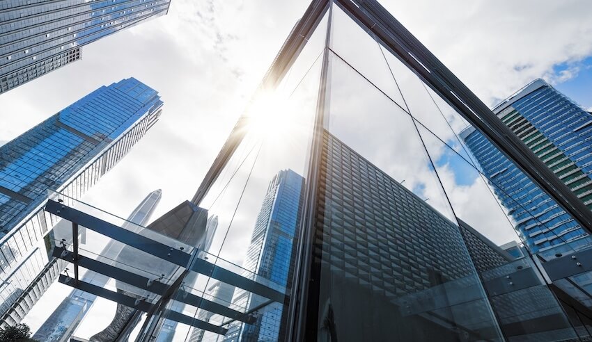 View looking up at modern glass skyscrapers with sunlight streaming through clouds, representing the public sector and urban development.