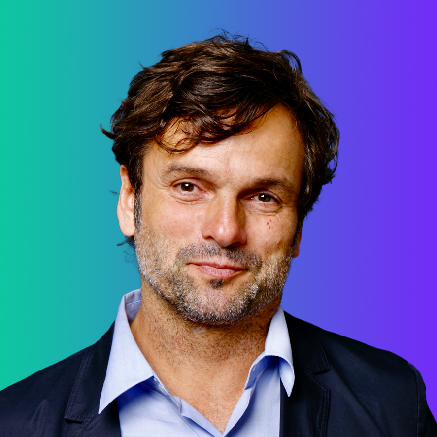 Portrait of a man with brown hair and beard, wearing a light blue shirt and dark blazer, in front of a green to blue gradient background.