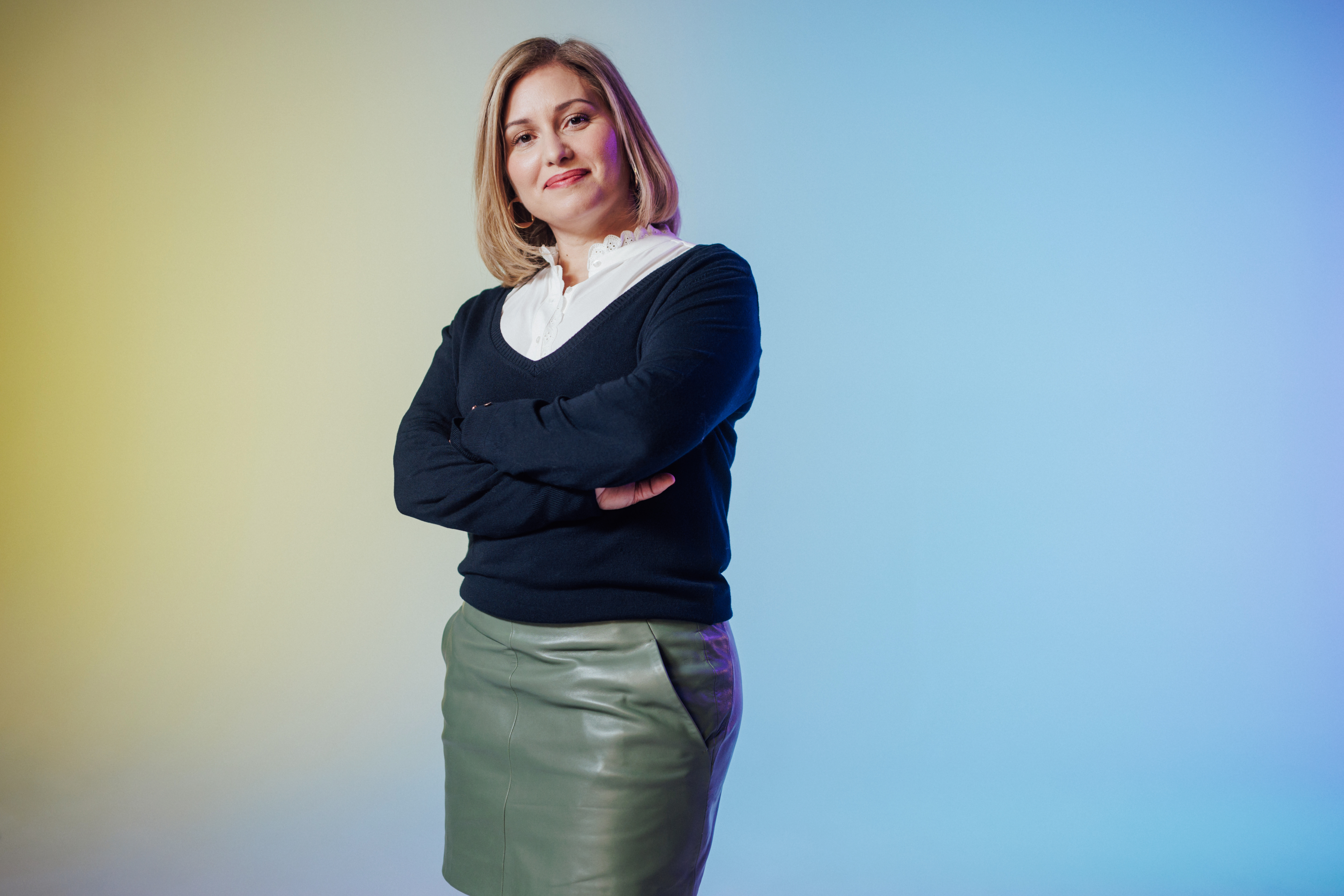 A woman standing with her arms crossed in a business outfit, smiling and posing confidently against a gradient blue and yellow background.