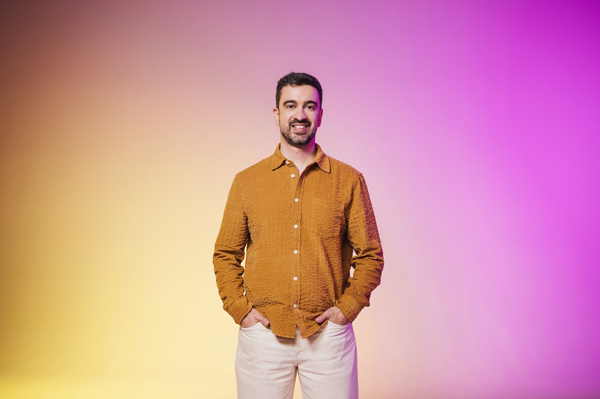 A man wearing a mustard shirt and white pants stands with his hands in his pockets, smiling, against a yellow and purple gradient background.