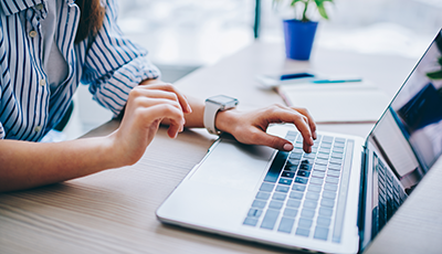 A person using a laptop computer at a desk, with a notepad and potted plant in the background.