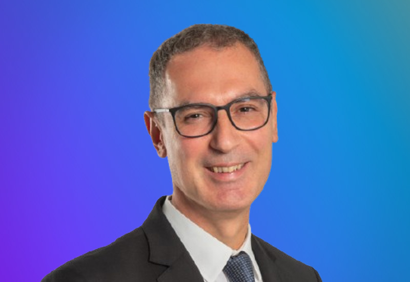 A man wearing a suit and glasses, smiling, in front of a blue and purple gradient background.