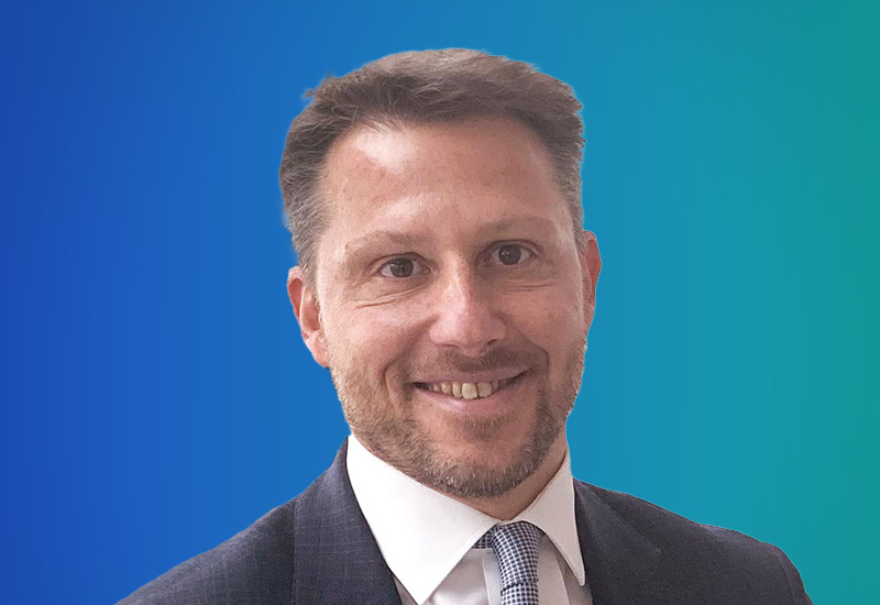 Professional headshot of a man in a business suit and tie, smiling, with a blue and teal gradient background.