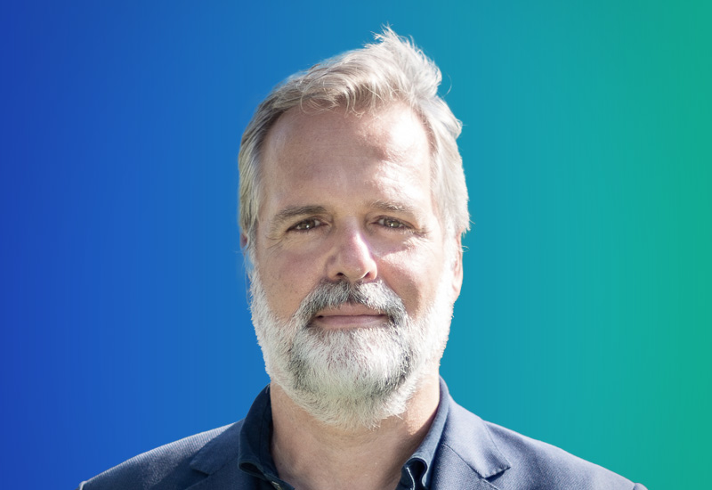 Professional headshot of a person with light hair and beard, wearing a dark suit jacket, in front of a blue-green gradient background.