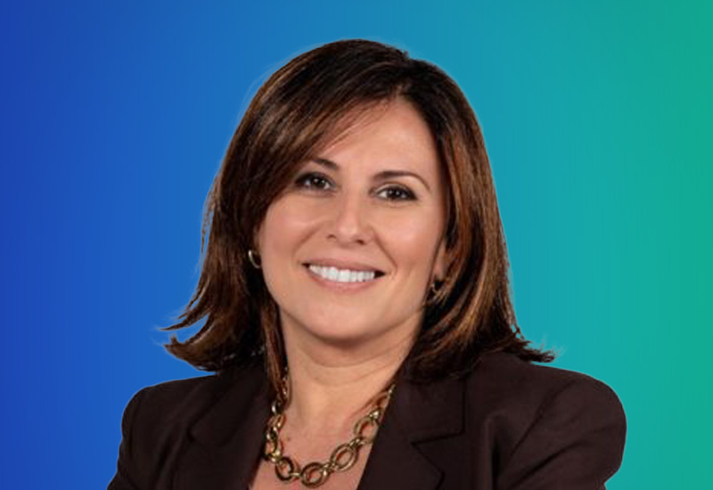 Professional headshot of a woman with shoulder-length brown hair, wearing a dark blazer and gold necklace, set against a blue and teal gradient background. This image is used for AWS Milano event promotions.