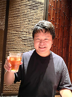 A smiling person holds up a glass of beverage while seated at a restaurant with textured and bamboo-patterned walls.