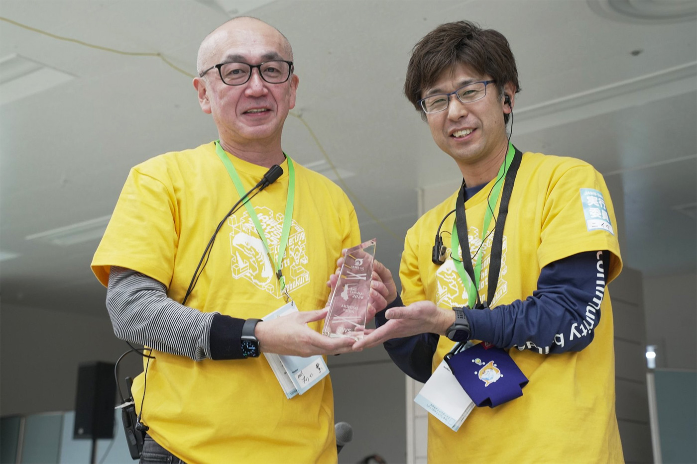 Two individuals in yellow shirts receiving and presenting an award at the AWS Samurai 2024 event.