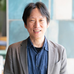 A casual portrait of a person wearing a gray jacket over a denim shirt, smiling in a softly lit setting.