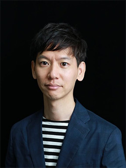 A portrait of a man wearing a dark blazer and a black-and-white striped shirt, posed in front of a black background.