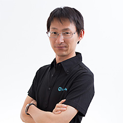A person with short dark hair and glasses is standing with arms crossed, wearing a black collared shirt, against a plain light background.