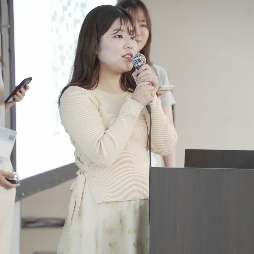A woman is speaking into a microphone at a podium, with other people and a presentation screen visible in the background.