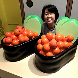 A person is smiling behind two oversized sushi-shaped objects filled with orange balls, each ball featuring a small face. The display is playful and cartoon-like.