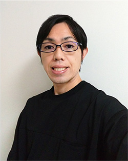 A portrait photo of a person with short dark hair, wearing glasses and a black shirt, standing in front of a plain light-colored background.