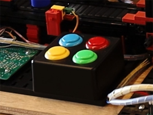 A close-up of a digital twin factory control panel featuring four multicolored control buttons (blue, red, yellow, and green) on a black base, surrounded by electronic components and wires.