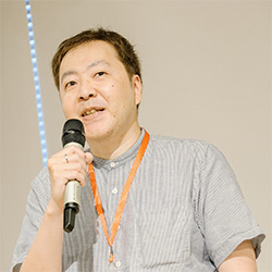 A man holding a microphone and speaking, wearing a striped shirt and an orange lanyard, photographed in profile against a plain background.
